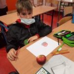 Fotografia przedstawia ucznia klasy 6a, Kamil Miński, który wykonuje pracę plastyczną. Chłopiec na kartce papieru ma narysowane jabłko, wykonane techniką cieniowania. Uczeń ubrany jest w czarną bluzę, a na piersi przyczepiony jest zielony emblemat. Na ławce można jeszcze zauważyć piórnik, kredki, blok i czerwony owoc.