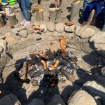 Na zdjęciu widać palące się ognisko otoczone polnymi kamieniami. Nad ogniem pieką się nadziane na patyki kiełbaski. Na dole zdjęcia widać cienie dzieci stojących przy ognisku.