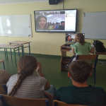 Zdjęcie zostało zrobione w sali lekcyjnej podczas quizu. Na zdjęciu znajdują się uczniowie Szkoły Podstawowej w Paczkowie wraz ze swoim nauczycielem przed dużym telewizorem, w którym widać uczniów wraz z nauczycielką z jednej ze szkół w Turcji. Witają się ze sobą.