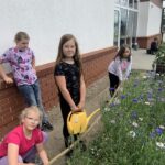 na zdjęciu widać cztery dziewczynki, które stoją obok poletka roślin. Dwie patrzą na rośliny, a dwie w obiektyw. Jedna z nich trzyma żółtą konewkę. Z tyłu za dziewczynkami widać chłopca zrywającego kwiaty.