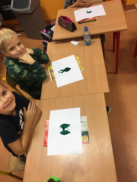 Chłopcy w kolorowych bluzach siedzą przy stolikach. Na stolikach leżą białe karty z odbitymi na nich zielonymi plamami.