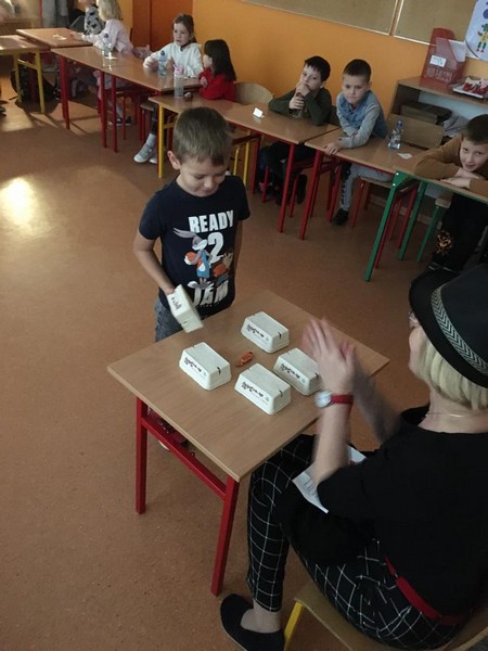 Chłopiec w czarnej koszulce z napisami podnosi pudełko, pod którym jest cukierek. Nauczycielka w czarnym stroju i kapeluszu bije brawo