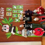 Siedmioro dzieci w opaskach z wizerunkiem pand stoi na tle dekoracji ściennych: wielki zielony bambus z uwieszoną na nim pandą. Obok powieszone są również prace plastyczne dzieci