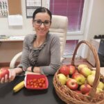 Fotografia przedstawia Panią Joannę, Naszego Sekretarza Szkoły. Pani Joanna uśmiecha się i trzyma w prawej dłoni zdrowe, czerwone i soczyste jabłko. Na biurku znajdują się pomidorki koktajlowe miseczce, banan oraz koszyk z jabłkami.  