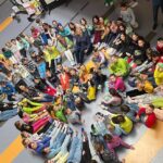 Zdjęcie wykonane z góry w holu szkoły. Dzieci siedzą na podłodze w kręgach i wystawiają kolorowe, różne skarpetki „nie do pary”.